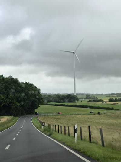 The image depicts a rural landscape in Weiler-la-Tour, Luxembourg, under a cloudy sky. A large wind turbine is visible in the distance, its blades blurred due to motion. In the foreground, a paved road curves to the left. To the left of the road, a dense line of dark green trees forms a hedge. To the right of the road, a grassy field is bordered by a wooden fence with vertical posts. There are at least two cars on the road, one dark-colored and one red, appearing to be in motion. The overall scene suggests a slightly overcast or rainy day, with water droplets visible on the lens or surface from which the photo was taken, particularly on the foliage. The environment is characterized by rolling hills, agricultural fields, and scattered trees.