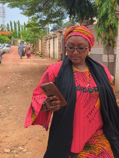 A woman is depicted standing outdoors on a sunny day in Unguwar Rimi, Nigeria. She is the primary subject, positioned slightly off-center and looking towards the viewer. She wears a vibrant, long-sleeved dress featuring a red background with intricate black and yellow patterns. A black headscarf or veil covers her head, and a black shawl is draped over her shoulders and upper body. She has glasses on, golden earrings, and two necklaces—one gold-toned and the other made of black and gold beads. Her hands are adorned with elaborate black henna designs, and she wears a simple bracelet on her right wrist. She holds a light brown smartphone in her left hand. Her expression is neutral.
The setting appears to be a semi-urban or rural area, characterized by a reddish-brown dirt path or road. Lush green trees and foliage are abundant, particularly to her left and in the background. In the middle distance along the path, two other individuals, also dressed in traditional long garments (one brown, one grey/blue), are walking away from the viewer. Behind them, buildings with light-colored pillars or columns are visible. Further in the background, parked vehicles can be seen, and a metal lattice tower structure is partially visible on the far left. The overall impression is daytime with clear weather. A small label on her dress, visible near her left hip, reads "DUTCH W".