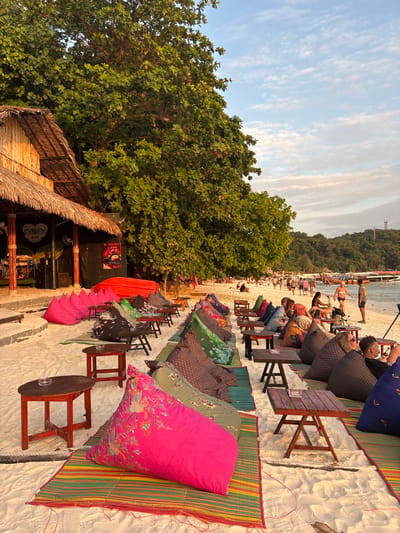 The image depicts a lively beach scene at Ban Ko Li Pe, Thailand, during golden hour. On the left, a wooden structure with a thatched roof, identified as "Monkey Bar," features an entrance flanked by bright pink bean bag chairs and a red inverted boat. Large green trees with thick foliage line the background behind the bar.
The foreground and mid-ground consist of rows of open-air seating arrangements on white sand. Each setup includes a colorful, patterned bean bag cushion placed on a striped mat, accompanied by a small, dark wooden table, some featuring ashtrays. Several individuals are seated on these bean bags, relaxing and engaging in casual interactions.
Towards the right and further down the beach, a larger group of people is seen walking along the water's edge. Some are in swimwear, others in casual attire, strolling along the shoreline. The calm, blue-green ocean in the background features several traditional longtail boats moored offshore. Beyond the beach and water, a lush, green hill rises, topped by a slender tower-like structure. The sky above is light blue with scattered cirrus clouds. The scene conveys a relaxed, evening atmosphere with warm, diffused lighting.