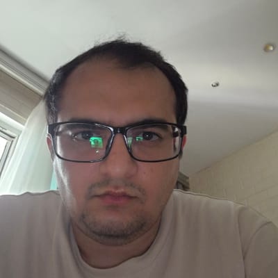 This media file is a close-up, rotated selfie of a person wearing black-rimmed glasses. The subject is a male with dark hair and a light beard. His eyes are dark, and reflections of a green light source are visible in his glasses. He is wearing a light-colored t-shirt. The background appears to be an indoor setting with a white ceiling and some textured wallpaper. There are also hints of a window or light fixture in the upper left corner. The lighting suggests it is daytime. The overall impression is casual. No text is visible in the image.