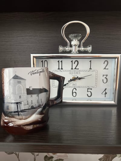 A close-up, eye-level shot captures a mug with a detailed image of a building and the word "Ventspils" printed on it, positioned to the left and slightly in front of a rectangular clock. The clock has a silver frame and displays the time as approximately 10:11. The mug has a brown and white marbled glaze and a dark brown handle. The clock face features large Arabic numerals for hours and smaller numerals in a rectangular inset. The background is a dark, textured wooden shelf. The location is not Sobishchytsi, Ukraine.