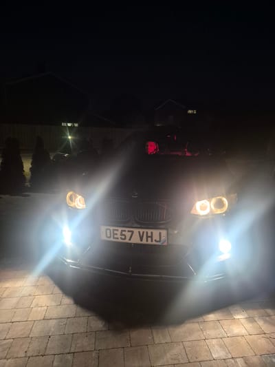The image depicts the front of a dark-colored car with its headlights on, illuminating a paved surface. The license plate reads "OE57 VHJ". In the background, there's a house and some vegetation. The scene appears to be at night, likely in a residential area within West Suffolk, United Kingdom.