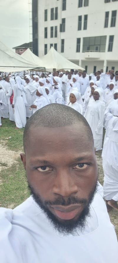 The image shows a man's face in the foreground, with a crowd of people wearing white clothing behind him. They appear to be outdoors, possibly in Anantigha, Nigeria, given the request for location. A building with many windows is visible in the background, along with a white tent. The man in the foreground is also wearing white clothing. There is grass and sand visible on the ground.