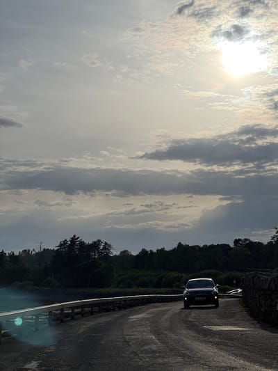The image depicts a dark-colored Kia Seltos driving down a road in Terhillion, Ireland. The sun is shining brightly in the sky, illuminating the clouds and casting a lens flare onto the road. The road is surrounded by trees and a stone wall. The car's headlights are on, suggesting it may be evening or a cloudy day. The overall mood of the image is serene and peaceful. The license plate of the car is visible and reads "20-TL-986".