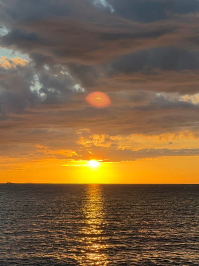 The image depicts a seascape at sunset, with the sun positioned low on the horizon, casting a warm orange glow across the sky and reflecting brilliantly on the rippling ocean surface. Dark, dramatic clouds with hints of orange and grey dominate the upper portion of the frame, partially obscuring the sun in one area and creating a lens flare effect. The sea occupies the lower half of the image, its dark, undulating surface shimmering with golden light from the sunset. A distant ship and a faint landmass are visible on the horizon, to the left of the setting sun. The overall scene is one of natural beauty and tranquility. This image was taken at Baan Plai Laem, Thailand.
