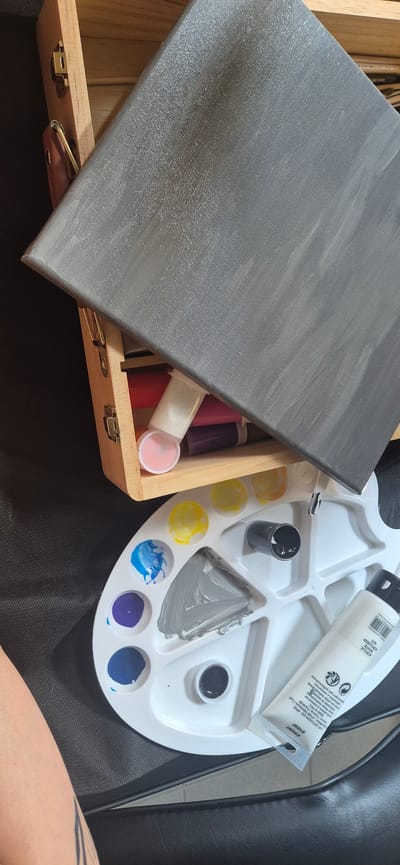 An overhead shot shows a white plastic paint palette filled with various colors of paint. A black paint tube is positioned in the top right of the palette. To the left of the palette is a wooden art supply box filled with paint tubes of various colors. A grey canvas is placed diagonally across the top of the art box. The background is a dark textured surface. There is a person's left arm and shoulder visible in the bottom left corner of the image, resting on the dark surface. The scene is indoors, with no indication of the specific location within Lozen, Bulgaria.