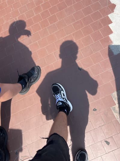 The image shows a ground-level view of a tiled surface, possibly a sidewalk or plaza, with two people's legs and shoes visible. The tiles are reddish-brown. The shadow of the individuals is cast onto the surface. One person is wearing black athletic shoes with a dark sock, and the other is wearing black and white sneakers with white laces and a dark sock. A third pair of dark sneakers is visible in the top right corner of the image. The person wearing the black and white shoes has a leg with visible skin and is wearing dark denim shorts with a frayed edge. The legs suggest a sunny day due to the shadows.
Given the reddish-brown tiles, the environment likely points to Bologna, Italy, which is well known for this distinctive architectural feature.