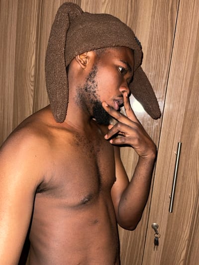 An adult male with dark skin, a beard, and a mustache is depicted indoors, bare-chested, wearing a brown textured hat with two long, floppy ear-like extensions. His head is turned to the right, and his gaze is directed downwards and rightward. His left hand is raised, with the index and middle fingers touching his upper lip and nose. The background features a light brown wooden cabinet or wardrobe with a vertical wood grain pattern. A silver-colored vertical handle is visible on the right side of the cabinet door, beneath which a lock with a set of two keys on a keyring is attached. The scene is illuminated by artificial light. The precise location within the city of Chikaji, Nigeria, cannot be ascertained from the visual evidence.