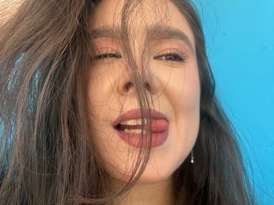 A close-up shot shows the face of a young woman against a bright blue background. Her dark hair is styled casually, with some strands falling across her face. She is looking towards the camera with a playful expression, her tongue sticking out slightly from her lips. Her eyes are partially closed, and she is wearing subtle makeup. A small silver earring dangles from her right ear. The lighting is bright, highlighting her features. The location is specified as Shulhynka, Ukraine.