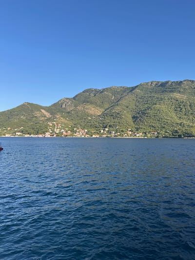 A wide-angle, eye-level shot captures a tranquil scene on the water with mountains and a village in the background. The foreground is dominated by deep blue water with gentle ripples reflecting the clear sky. In the middle ground, a sun-drenched village with red-tiled roofs lines the shore, nestled at the base of lush green mountains. The mountains, covered in dense foliage and dotted with scattered buildings, rise steeply into a bright blue sky. The overall impression is one of peaceful coexistence between human habitation and the natural landscape.