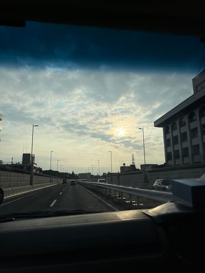 The image shows a view from inside a vehicle traveling on a multi-lane highway, likely during daylight hours as indicated by the bright sky and visible sun. The highway is divided by lane markings, and there is a metal guardrail along the right side of the road. Several vehicles are visible on the highway, including a white car in the far right lane and other cars further ahead. On the left side of the highway, there are sound barriers and some buildings in the background, including a multi-story building with many windows. Streetlights are also visible along the sides of the highway. The sky is partly cloudy with the sun visible through the clouds, creating a bright spot in the upper center of the image. A dark tinted band, likely the top of the vehicle's windshield, runs across the top of the image. The overall scene suggests a typical urban or suburban highway environment in Japan, possibly Itabashi, given the architectural style of the buildings and the presence of the guardrails and sound barriers.