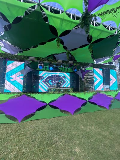 An outdoor stage setup is depicted from a frontal, slightly low-angle perspective. The scene is set in Noumea, France. A large, complex canopy structure, supported by metal trusses, dominates the upper portion of the frame, featuring numerous interconnected square fabric panels in shades of lime green, dark green, and light grey/white. Artificial green foliage is suspended from the canopy and draped along the stage's upper edge.
The stage itself occupies the mid-ground, centered with a performer, a male DJ in dark clothing, standing behind a console. He is actively operating professional DJ equipment, including turntables/CDJs and a mixer. Behind the DJ, a central LED screen displays a dynamic blue and white geometric pattern with partial text visible ("_TTR"). Flanking this central screen are two additional large LED screens, each exhibiting a repetitive blue and white chevron pattern. Black line array speakers are mounted vertically on either side of the central screen. Multiple blue-lit stage lights are positioned above the central screen, casting illumination forward.
A decorative barrier, similar in construction to the overhead canopy, extends across the foreground at the base of the stage. This barrier features alternating vibrant purple and green square fabric panels. The ground in front of the barrier is a meticulously manicured, short-cut grassy area. A clear blue sky is visible through gaps in the overhead canopy.