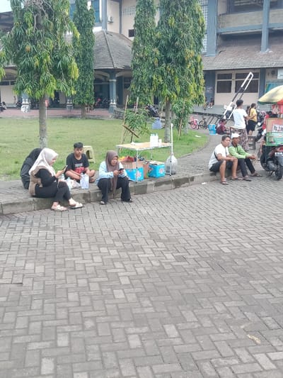 A street scene shows several people sitting on the edge of a curb. Three people are seated together looking at their phones. Another person is seated alone. Next to them is a vendor table with goods for sale. Further down the curb, two men sit and chat, with a motorcycle nearby. In the background, there is a building with tall trees lining the front. People, bicycles and a food cart are visible in the distance. The foreground is filled with a gray brick paved area.