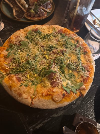 The scene depicts a dining table, likely within a restaurant in San Agustín, Mexico, though the precise establishment is not discernible from the image alone, beyond the branding "Agos Bon" visible on a glass.

In the foreground and center, a large, round, cooked pizza dominates, featuring a thick, golden-brown crust. Its surface is generously covered with melted cheese, scattered green arugula leaves, numerous light green, shelled pistachios, and thin, reddish-pink slices of cured meat. Additional shredded white cheese is visible over the toppings.

In the upper-left background, a dark green plate holds two separate dishes: one with two bone-in cuts of meat, and another with multiple small, round items resembling tacos or tostadas, topped with various ingredients.

To the right of the pizza, a tall, clear glass contains a dark beverage with ice and a beige straw. Further right, an empty clear glass displays the "Agos Bon" branding. Behind these glasses, a small, light-colored ceramic plate holds a piece of bread and cutlery.

In the lower-right foreground, a small, dark bowl contains a viscous dark liquid, with a metal spoon resting within it. The items are arranged on a dark, textured tabletop, possibly marble or stone, under dim, ambient lighting. No individuals are present in the frame.