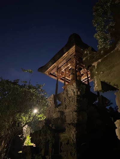 The image depicts a night scene in Ubud, Indonesia, showcasing a traditional Balinese structure, likely a temple or shrine. The structure has an ornate, multi-tiered roof with intricate carvings and wooden beams. It's situated within a stone enclosure adorned with similar carvings. A tree with dense foliage is visible in the foreground, illuminated by a nearby light source. The background reveals a dark blue night sky.