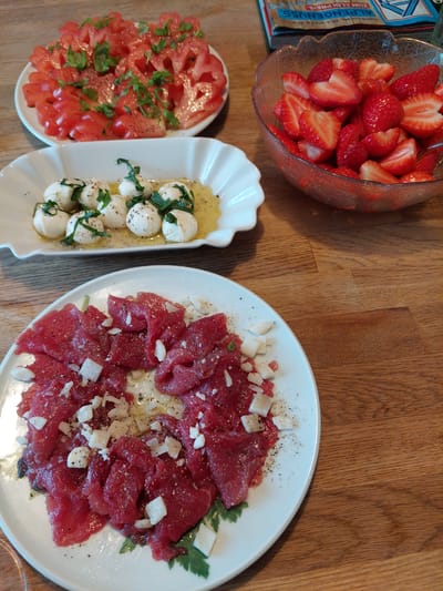 A top-down, natural light shot shows a wooden table set with an array of dishes. A glass bowl filled with sliced strawberries is in the upper left. Below the strawberries, two white serving dishes hold chopped tomatoes with basil and mozzarella balls with basil and olive oil, respectively. To the right, a large white plate is covered with thinly sliced raw meat, topped with diced white cheese or onions, and scattered with pepper. A box of Alpengenuss chocolate is partially visible. The location is indoors on a wooden table. The city of Ganderkesee, Germany, is not discernible from the visual information.