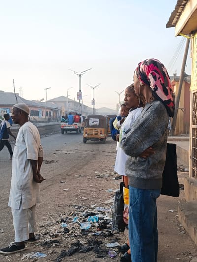 An outdoor scene shows three people standing on the side of a street with trash scattered on the ground. A man wearing a white cap and robe stands closest to the foreground, with his back partially turned. A woman with a child stands beside him, and a woman with a pink and black headscarf stands nearest to the viewer, her back turned. In the background, motorized rickshaws and a vehicle with people in the back move along the street. Overhead street lights line the road. Buildings are seen along the horizon.