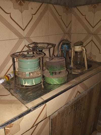 This image depicts an indoor utility area or kitchen within Nyansiongo ward, Kenya. The background wall is covered with light brown ceramic tiles featuring a repeating pattern of dark brown geometric lines and stylized floral motifs. A soiled metal or tiled countertop occupies the middle ground, with a visible metal sink basin located on the right.
Arranged on the countertop are several vintage items. From left to right, a prominent, heavily rusted dark metal framed kerosene or paraffin stove with a teal-green cylindrical fuel tank is visible. Next to it, a smaller, similarly rusted stove with a dark metal top and a green base is present. Partially obscured behind the leftmost stove is the rusted metal frame of another stove burner. Inside the sink basin, two vintage kerosene lanterns are placed. The left lantern features a blue top and a clear glass globe, while the right lantern is off-white with a clear glass globe displaying an "ANCHOR" brand label. On the far left of the countertop, a small yellow and white squeeze bottle with a white cap rests horizontally.
All metal objects show significant rust, dirt, and general wear. The countertop surface exhibits grime and reflective properties. Below the countertop, additional patterned tiles match the wall design, alongside a section of dark wooden paneling, possibly a cabinet door. No individuals are present in the scene.