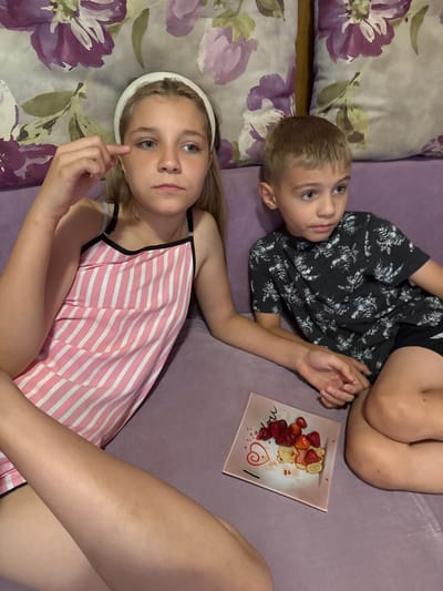 The image shows two children lying on a purple couch with floral patterned pillows. The boy, on the right, is wearing a dark t-shirt with a white floral pattern and black shorts. He is lying on his back, looking up and to the left. The girl, on the left, is wearing a pink and white striped tank top. She is lying on her back, looking directly at the camera with her right index finger raised to her cheek. A small, square pink plate with strawberries and a teddy bear design is placed on the couch between the children. The lighting suggests it is indoors, possibly during the day. The overall mood appears relaxed and casual. No visible text is present in the image.