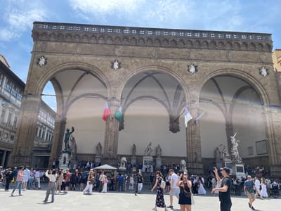 The image shows the Loggia dei Lanzi, an arcaded building in Piazza della Signoria, Florence, Italy. The Loggia houses sculptures. A large crowd of people is gathered in front of the Loggia, some taking photographs. Flags hang from the arches of the Loggia. The sky is blue with some clouds.