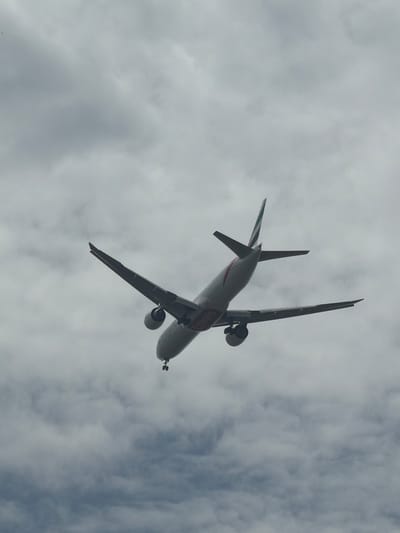 An underside view of a commercial airplane is captured in flight against a cloudy sky. The aircraft, a narrow-body jet with twin engines mounted under the wings, is seen from below, with its landing gear extended. The tail of the plane features a vertical stabilizer with a distinctive green and red striped livery, and red and white banding is visible along the fuselage. The words "Emirates" are partially visible on the side of the plane. The surrounding environment is entirely obscured by a dense, overcast sky composed of gray clouds. There is no indication of the city of Delhi, India, or any other specific location, people, or activities in this image.