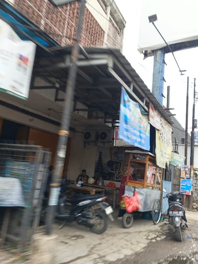 A street scene shows a food vendor cart under a covered area with colorful banners. A woman stands inside the cart. A table sits nearby with a man seated. Two motorcycles are parked close to the cart and table. The buildings appear to be in disrepair. Overhead, a large billboard is visible. Utility poles line the street's edge. The overall atmosphere indicates a busy, though somewhat run-down, commercial area.