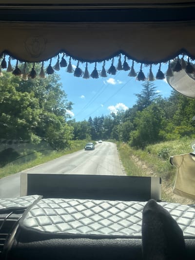 The image, taken from inside a vehicle in Topolovgrad, Bulgaria, captures a view through the windshield. A decorative trim with numerous tassels hangs across the top of the windshield. The road ahead is paved and curves to the left, bordered by lush green trees and vegetation. Two cars are visible on the road in the distance. The sky is blue with scattered white clouds, and power lines run overhead. In the foreground, the interior of the vehicle is visible, with a quilted dashboard and what appears to be a black sock or fabric in the lower right corner. A beige garment is also hanging from the right side of the frame.