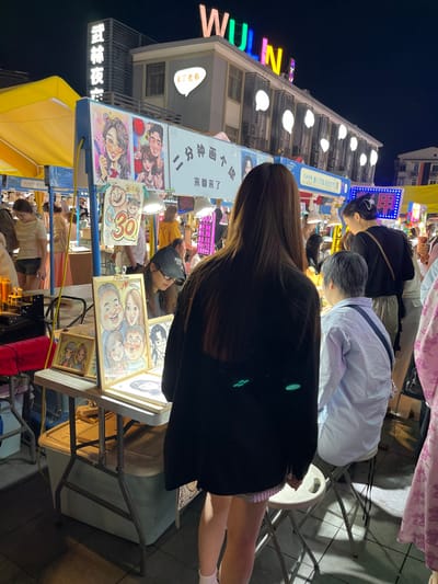 The image shows a street scene in Hang Zhou Shi, China, at coordinates 30.264 latitude and 120.156 longitude. It's a night market with various vendors, including a caricature artist who is drawing a portrait of a man. The artist is sitting at a table with other drawings displayed. A woman with long dark hair is standing by the table, seemingly observing the artist's work. Behind her is a man wearing a light blue shirt, also observing the process. The scene is dimly lit, and the atmosphere is casual and relaxed. The building in the background has a sign that reads "WULN". There are many other people walking around in the night market, but they are not the focus of the image. The focus is on the artist, the woman, and the man.