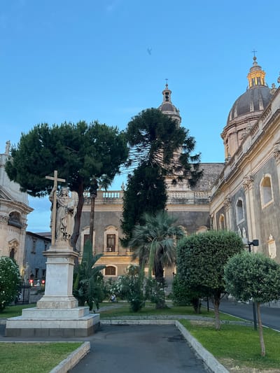 The image depicts a twilight scene in Piazza Duomo, Catania, Italy, featuring the Catania Cathedral. In the foreground, a white marble statue of a woman holding a cross and a book stands on a tiered pedestal. Behind the statue, a large pine tree and a palm tree are visible, with manicured bushes and green grass in the immediate vicinity. The Catania Cathedral's imposing facade and dome dominate the background, with architectural details such as arched windows and ornate carvings visible. The sky is a clear blue, transitioning to dusk. A faint bird is captured in flight against the sky. The lighting suggests a late afternoon or early evening setting, with the warm glow from the cathedral dome indicating artificial illumination beginning to activate. A person is seated in the background, in the grassy area. The overall atmosphere is serene and historic, characteristic of a European piazza.