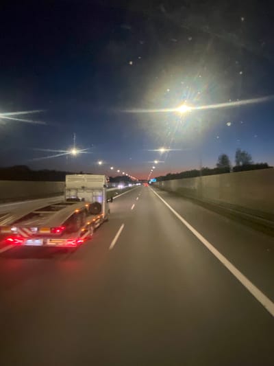 The image depicts a night scene on a highway, likely in the vicinity of Podłęże, Poland, as indicated by metadata. The perspective is from within a vehicle, looking forward. The road surface is asphalt with a white lane marking visible. On the left side of the road, a white trailer truck with a flatbed is visible from the rear, its brake lights and taillights illuminated red. The truck appears to be transporting something on its trailer, though the cargo is not clearly discernible.
The sky is a deep blue transitioning to a dark twilight, with the silhouette of trees visible in the distance on the right side, behind a sound barrier wall. Streetlights are present along the side of the highway, casting a bright glow that creates lens flares in the image. The overall impression is of travel on a highway at dusk or night. There are no people clearly visible in the image. The environment is an open highway, with traffic lights and distant vehicle lights suggesting other vehicles are present on the road ahead.