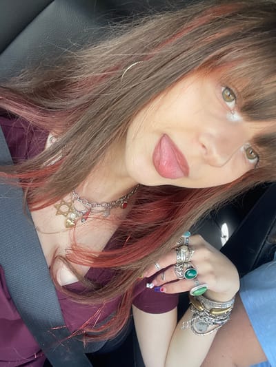 The image depicts a female individual, observed from the chest up, situated within the interior of a vehicle. Her head is tilted, and her tongue is extended from her mouth. She has long, dark brown hair with red or auburn highlights and bangs that frame her forehead. Her eyes are light brown or hazel. She is wearing a maroon-colored V-neck garment. A dark grey seatbelt is visible across her left shoulder and chest.
Around her neck, multiple layered necklaces are present; one prominent silver-colored chain features a large gold-colored Star of David, a gold-colored heart, and a small red heart charm. A silver-colored hoop earring is visible in her left ear. Her left hand is prominently displayed, adorned with numerous silver-colored rings, some incorporating large stones such as green, white/opalescent, and light blue. Her left wrist is heavily accessorized with multiple silver-colored bracelets. Her fingernails are painted with multi-colored polish, including a red heart design on one nail.
The background consists of dark, possibly black, vehicle upholstery. A partial view of a light blue garment or fabric is visible on the right side of the frame, suggesting another occupant or object. There are no identifiable visual elements within the image to confirm the specific location as Kfar Saba, Israel.