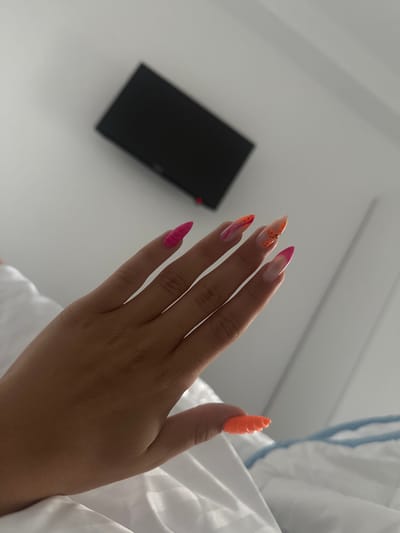 The image shows a close-up of a person's left hand with long, stiletto-shaped acrylic nails painted in various vibrant colors and designs. The nails are decorated with pink, orange, and yellow hues, some featuring abstract patterns and speckles. The hand is resting on a white, possibly satin or silk, fabric, likely bedding. In the background, a portion of a black, rectangular object, possibly a TV screen, is mounted on a light-colored wall. The overall setting appears to be indoors, in what could be a bedroom or hotel room, with a bright, diffused light source. There is no specific indication of the location being Stațiunea Zoologică Marină Agigea, Romania.