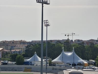 The image shows a scene in Figueira da Foz, Portugal. Two white circus tents are visible with trailers and vehicles surrounding them. In the background, a cityscape with multiple buildings and trees stretches across the horizon. Two tall stadium light poles stand prominently in the foreground. Construction cranes are visible among the buildings in the background. A sports stadium is suggested by the presence of the light poles and the field area visible at the bottom of the frame.