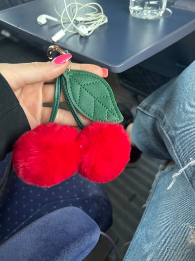 A person's hand with bright pink fingernails holds a keychain with two fluffy red cherry charms and a green leaf charm. The background shows a blue seat with a dotted pattern, a grey and blue tray table with headphones and a water bottle on it, and a person wearing ripped blue jeans. The scene is likely in Kaspichan, Bulgaria, possibly on public transportation or in a waiting area.