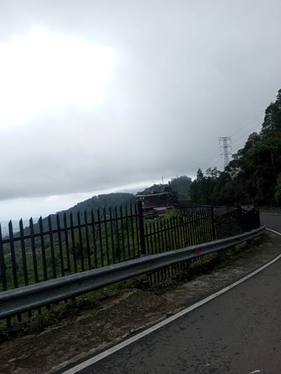 The image displays a mountainous landscape in Bangtsho, Bhutan, under a heavily overcast sky. A dark, possibly damp, asphalt road with a solid white edge line occupies the lower right foreground, curving gently to the right. Parallel to the road, a silver-colored metal guardrail with at least two visible red circular reflectors is present. Immediately beyond the guardrail, a black metal fence, composed of vertical pickets with pointed tops, extends across the mid-ground, securing the edge of a steep, densely vegetated hillside. In the mid-distance on the left, an expanse of hills and a valley are partially obscured by atmospheric haze or low-hanging clouds, revealing glimpses of distant urban elements or structures. On the central mid-ground hillside, several multi-story buildings with reddish-brown sloped roofs are discernible amidst the green foliage. To the upper right, a large metallic lattice power transmission tower stands prominently on a higher, thickly forested slope, with power lines extending from it. The overall environment is characterized by steep, green, mountainous terrain under a uniform grey sky. No individuals, vehicles, or specific actions are observed within the frame.