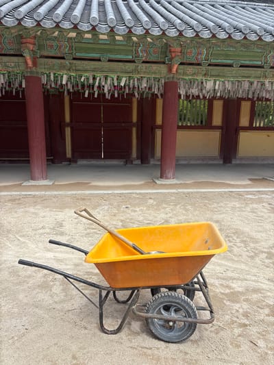 The image shows an orange wheelbarrow with a shovel inside, parked on a sandy ground in front of a traditional Korean building. The building features a tiled roof, ornate wooden eaves decorated with colorful paintings, and red pillars. Many small white papers are hanging from the eaves, possibly prayer tags or wishes. The building's walls are made of dark red wood, with some windows or openings revealing greenery behind them. The ground in the foreground is a mix of sand and dirt, with some patches of light gray concrete. The overall scene suggests an outdoor area, possibly a temple or historical site undergoing maintenance or renovation in Gyeongju-si, South Korea. There are no people or animals visible in the image.