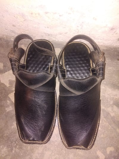 A pair of black leather Peshawari chappals are displayed on a concrete floor. The chappals are positioned side-by-side, with the toe portions facing the viewer. They feature a traditional design with a broad strap across the vamp and a narrower strap that goes over the instep and connects to a heel strap. The soles appear to be made of leather with a visible stitching detail around the edges. The background consists of a textured, light-colored wall with subtle variations in tone. The lighting suggests an indoor setting, possibly during the evening or in a dimly lit area. The image was captured in Haveli Jhangranwali, Pakistan.