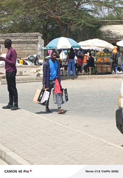 The image captures an outdoor scene, likely a street or marketplace, under bright daylight. The ground in the foreground is paved with light-colored square or rectangular blocks, resembling cobblestones or large tiles.
In the left foreground, a man stands on a raised curb or sidewalk, facing partially away from the camera. He wears a long-sleeved maroon or dark red shirt, dark trousers, and dark closed shoes. His left arm is bent, and he appears to be holding a dark rectangular object, possibly a smartphone, in his hands, looking down at it.
In the mid-ground, a second man walks across the paved road, moving from left to right. He is dressed in a light blue t-shirt layered under an open dark blue or grey jacket, grey trousers, and open-toed sandals or flip-flops. He carries several shopping bags in both hands; two black bags with white text/logos are visible in his left hand, and a brown paper bag is in his right hand, along with another dark bag. A distinctive red, tassel-like object hangs from his left hip area.
The background reveals a bustling market area. On the left side, behind the first man, there's a low, rough-hewn stone wall, behind which appear to be some discarded items or debris, including black trash bags and other objects on the ground. Further into the background, beneath the canopy of trees with sparse foliage, are several market stalls. Two large umbrellas provide shade over these stalls: one is light blue and white (with some illegible green text), and the other is white. Under these umbrellas, several people are gathered around the stalls. One stall prominently displays fresh fruits, including several large watermelons and a pile of bright orange fruits, possibly oranges or mandarins. Other figures are visible interacting or standing near these stalls; one person is seen in blue pants and a dark top, and another person in a red top/dress is also present. A person is sitting on a stool under the white umbrella.
The lighting indicates a sunny day, with clear shadows cast by the people and objects, suggesting it is likely mid-day. On the far right edge of the frame, the front corner of a white vehicle is partially visible.
Technical details about the image capture are noted at the bottom of the frame: "CAMON 40 Pro" (bottom left) and "96mm f/1.8 1/1499s ISO52" (bottom right).
