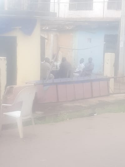 The image depicts a group of men sitting and conversing outside a building. The setting appears to be in a residential or commercial area, possibly a shopfront, with a light yellow and blue facade. A white plastic chair is prominently visible in the foreground. In the background, a multi-story building with windows can be seen. The overall atmosphere is casual, with the men engaged in conversation. The scene suggests a typical day in the city of Enugu, Nigeria, where such informal gatherings are common.