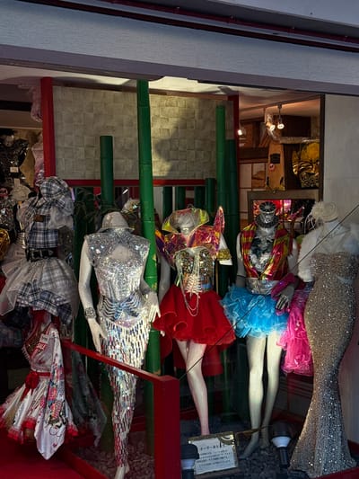 This image captures a vibrant and densely packed display of elaboratorate costumes, likely from a specialized shop in Shibuya, Japan. The scene features multiple mannequins, arranged in layers, showcasing a diverse range of flamboyant attire.
From top to bottom, the mannequins are dressed in distinct styles. One wears a sparkling gold sequined mermaid-style gown, accented with a white feathered boa and a decorative fascinator. Another nearby mannequin sports a bright pink ruffled dress. Below them, a mannequin features a metallic iridescent top paired with a voluminous blue tulle skirt and fishnet stockings. Further down, a mannequin is dressed in a bold red ruffled skirt and a shiny, possibly holographic, top. A prominent mannequin in the mid-ground stands out with a dazzling silver sequined top and geometric mirrored pants, giving a futuristic or disco-inspired look. At the bottom, mannequins display a plaid outfit consisting of a mini-skirt and top, and a traditional white Japanese-style robe or kimono adorned with red ties and floral patterns.
The display is structured within a red framework, complemented by vertical green poles resembling bamboo. The background reveals tiled walls and an interior space with warm lighting, suggesting an indoor setting or a shop window display at night.
Visible text on a sign in Japanese reads: "オーダー承ります" (Custom orders accepted), "衣装舞台衣装・コスプレ" (Costumes, stage costumes, cosplay), and "インやサイズも自由にオーダーできます。" (Designs and sizes can be freely ordered). Price tags include "6980" (likely Japanese Yen) and "SALE." The letter "S" is also visible, possibly indicating a size. The overall impression is that of a bustling shop specializing in unique performance wear and custom costume design.
