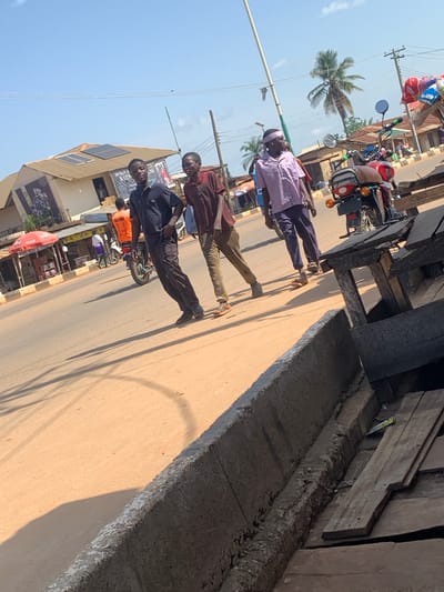 The image depicts a street scene in Kafanchan, Nigeria. Three young men are walking on a dirt road under a clear blue sky with scattered clouds. The man on the left wears a dark polo shirt and dark trousers. The middle man wears a brown collared shirt and khaki trousers. The man on the right is dressed in a pink and white checkered shirt and dark trousers, with a light-colored bandana on his head. In the background, buildings with tiled roofs, solar panels, and awnings line the street. A red umbrella marks a stall. A red motorcycle is parked to the right, with various goods displayed on a stall made of wood. A palm tree is visible in the distance. The ground is a light brown dirt. The overall scene appears to be a typical day on a street in a Nigerian town.