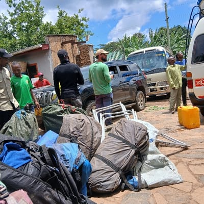 The image depicts an outdoor staging area in Mweka, Tanzania, under a partially cloudy sky. Six adult males are present, along with three vehicles and numerous pieces of luggage and equipment.

In the foreground, a cluster of large, soft bags or duffels in various colors (olive green, dark brown, black, blue, and light grey) are piled on a reddish-brown paver and dirt surface. Several disassembled or folded metal-framed chairs are also visible on the ground. To the immediate right, a yellow plastic jerrycan rests on the ground next to a white van.

From left to right, the individuals and vehicles are arranged as follows:
*   A man in a light-colored shirt and black cap stands on the far left, partially obscured.
*   Next to him, a man in a green t-shirt stands.
*   Behind them and to the left, a portion of a simple building with a corrugated iron roof and stone-clad facade is visible. A man in a red shirt is partially visible inside a dark grey SUV, which is parked facing the foreground. This SUV, likely a Toyota Land Cruiser Prado, is dark grey.
*   A man in a black long-sleeved shirt and black cap stands with his back to the camera, facing the dark grey SUV.
*   To his right, a man in a green t-shirt, beige wide-brimmed hat, and sunglasses stands near the open passenger door of the dark grey SUV, looking towards its interior or side.
*   Behind the dark grey SUV, a beige van or minibus, resembling a Toyota Hiace, is parked.
*   To the right of the beige van, a man in a light green shirt and light-colored pants stands, looking towards a white van or minibus, also resembling a Toyota Hiace, which is partially visible on the far right.
*   A seventh man in a blue shirt is visible further back, near the white van.

Lush green trees and vegetation are present in the background, particularly on the right side. The scene suggests preparations for travel or an expedition, with equipment being handled or organized.