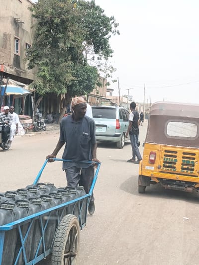 The image presents a daylight street scene in Hotoro South, Nigeria. In the immediate foreground, an older male, wearing a dark blue collared shirt, dark trousers, and a patterned brown and white cap, pushes a two-wheeled, blue-framed cart. The cart is loaded with approximately sixteen black plastic jerry cans, arranged in four rows. The man is facing towards the right side of the frame, moving forward.
To the right of the man and slightly behind him, the rear of a yellow three-wheeled auto-rickshaw (keke napep) with a brown fabric roof is visible, oriented away from the viewer. It features a single red taillight on the left rear and a louvered engine cover. Further back on the street, a light-colored minivan, possibly silver or light blue, is either parked or moving, displaying a license plate reading "BW 374KA". A younger male, dressed in a white t-shirt, dark vest, and dark trousers, is walking on the street behind the minivan.
On the left side of the frame, two individuals are seated on a motor-scooter; the rider wears a plaid shirt, and the passenger a white garment. Behind them, the roadside is lined with various commercial structures and stalls, including one with a blue awning. A large, leafy green tree stands prominently on the left, partially obscuring a multi-story building constructed from unrendered concrete blocks, which has visible windows. The street surface is paved. Utility poles and overhead lines are visible in the distant background under a bright, overcast sky.
