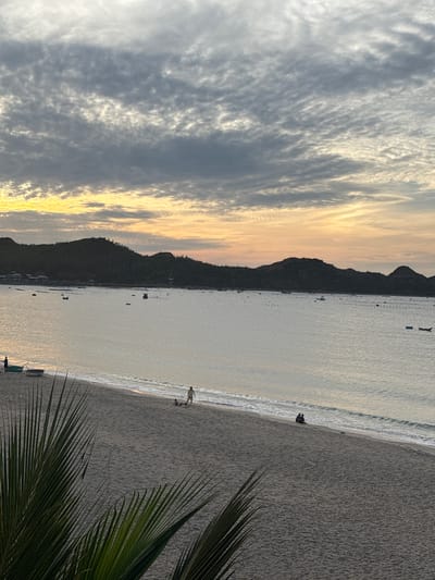 A wide-angle, low-light, eye-level shot captures a serene beach scene at dusk in Lệ Uyên, Vietnam. The sky is dominated by dramatic, textured clouds in shades of gray and white, with a warm yellow-orange glow on the horizon, suggesting the last moments of sunset. Below the sky, dark, silhouetted hills form a distant coastline. The calm sea reflects the soft light, dotted with numerous small boats and what appear to be aquaculture cages. A sandy beach occupies the foreground, with gentle waves lapping at the shore. On the beach, three individuals are visible: one person stands near two small boats on the sand, another person walks along the water's edge, and two people sit together near the waterline. Lush green palm fronds are in the lower left corner, framing the scene. The overall atmosphere is peaceful and tranquil.