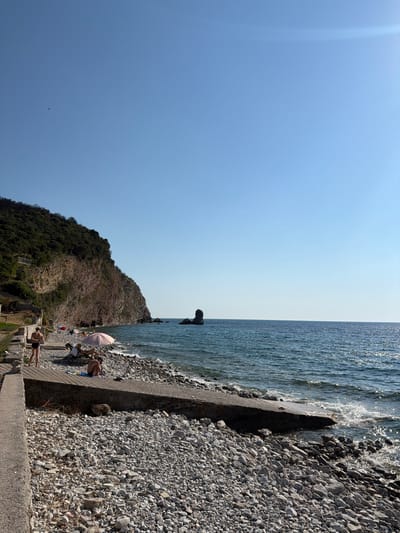 The image depicts a sunny day at a rocky beach in Golubovići, Montenegro. The scene is split between the bright blue sky on the left and the shimmering ocean on the right. A rocky shoreline is visible, with waves gently lapping against it.
Several people are present on the beach, enjoying the weather. Some are sunbathing on chairs, while others are casually lounging on the rocky shore. One person is lying down on a towel, another is sitting in a beach chair under a pink umbrella, and a third is standing, seemingly looking out at the sea. In the foreground, a person is doing a yoga pose on the edge of the beach.
The background features a lush, green, and rocky hillside that slopes down towards the sea, adding a natural element to the coastal scene. The water is clear and reflects the sunlight, creating a sparkling effect. The overall atmosphere appears relaxed and tranquil, suggesting a peaceful day at the beach. The time of day appears to be midday, given the bright sunlight and the shadows cast.