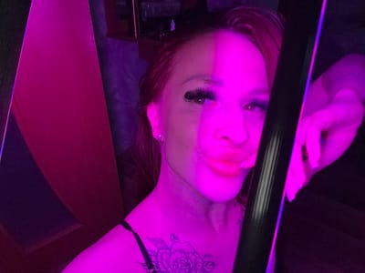 The image shows a person from an upside-down perspective, likely taken with a camera or phone held in that orientation. The person has light skin, red hair, and is wearing what seems to be false eyelashes and lipstick. There's a tattoo visible on the upper chest or shoulder. The lighting is primarily purple or magenta. In the background, there is what appears to be a door and a dark object or structure. There's also an elongated, black object in the foreground. The location is not discernible as Simferopol, Ukraine.