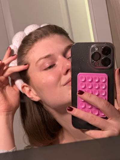 The image depicts a female subject, partially visible from the chest up, reflected in a mirror within an unidentifiable indoor setting in the city of Бакча-Сарай, Russia. She has light brown hair held back by a white, segmented, soft-textured headband. A small, clear liquid droplet is visible on her right cheek. Her fingernails are painted dark red. The subject holds a dark-colored smartphone, featuring a prominent triple-lens camera array and housed in a clear, glitter-infused case. Attached to the back of the phone is a bright pink, rectangular, multi-bubble silicone accessory. Her right hand is positioned around the phone, while her left hand gently touches the headband near her left temple. The background reflection shows indistinct, light-colored surfaces typical of an interior room.