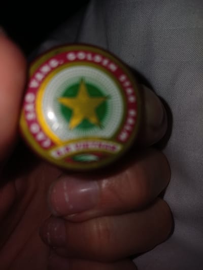 The image shows a close-up of a person's hand holding a circular object with a yellow star inside a green circle, surrounded by red and white text. The object appears to be a balm or ointment tin. The background is blurred but seems to be fabric, possibly clothing. The lighting is dim. There is no discernible location in the city of Middlesbrough, United Kingdom.