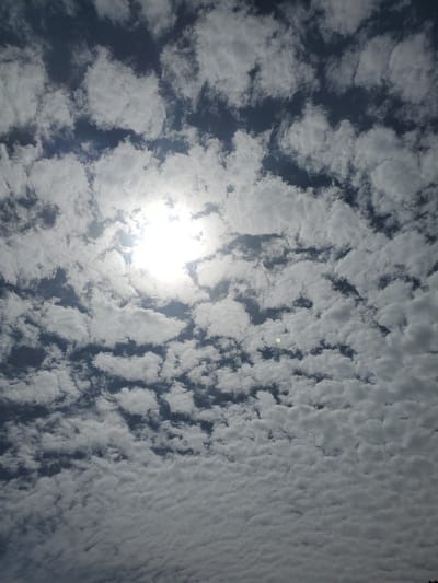 The image depicts a sky filled with a mix of dark blue and white clouds, with the sun prominently visible in the upper left quadrant, partially obscured by clouds. The clouds appear to be cumulus and stratocumulus formations, creating a textured, dappled effect across the sky. Sunlight filters through the clouds, illuminating some of them and casting shadows on others. The overall impression is of a bright, partly cloudy day. There are no people or man-made objects visible, suggesting the photograph was taken outdoors, looking upwards. The scene is natural and evokes a sense of calm or perhaps a dramatic sky. No text is visible in the image. The location context of Kabwir, Nigeria, suggests a tropical or subtropical climate, which is consistent with the type of cloud formations and the intensity of the sunlight. The time of day appears to be mid-morning or early afternoon, given the sun's position and brightness.