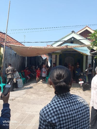 The image shows a gathering of people in an outdoor space, likely a courtyard or street, in Anjatan, Indonesia. There is a large awning stretched across the space, providing shade from the sun. Several people are sitting on chairs, while others are standing and interacting. A few children are playing in the space. There is a motorbike parked on the right side of the image, which is also in the shade of the awning. The environment is generally clean and well-maintained, with a paved surface and some greenery in the background.